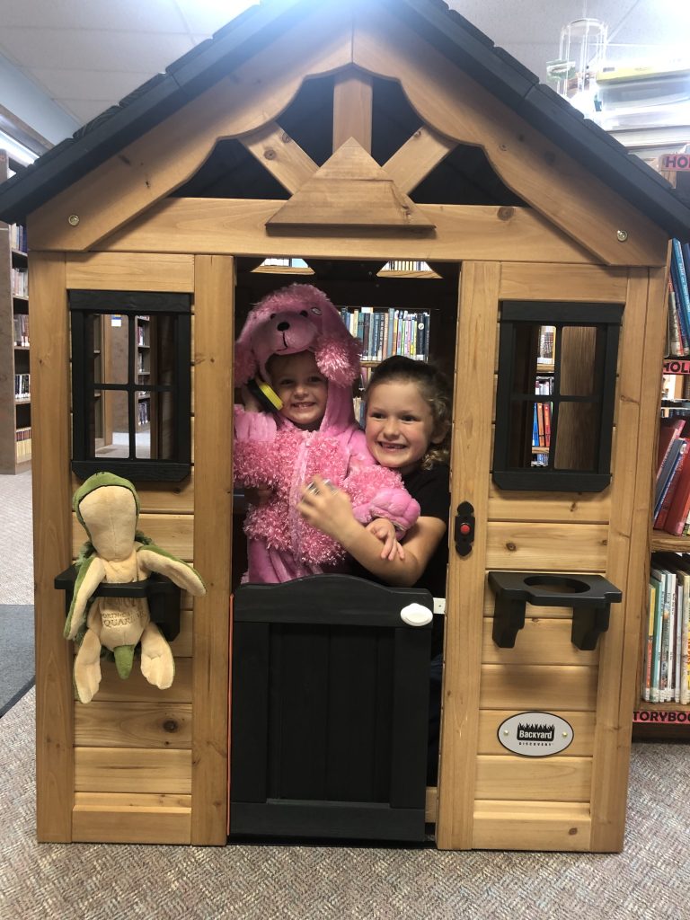 An image of a small playhouse with two children inside. The house looks like a wood cabin with a half door in front for access.