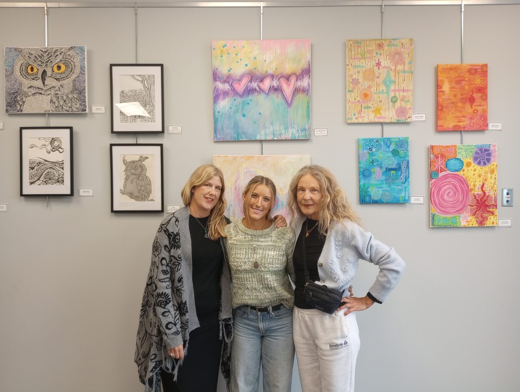 Photo of three women in front of the art wall display space, with multiple works in different mediums. The women are implied to be the artists responsible for the works. 