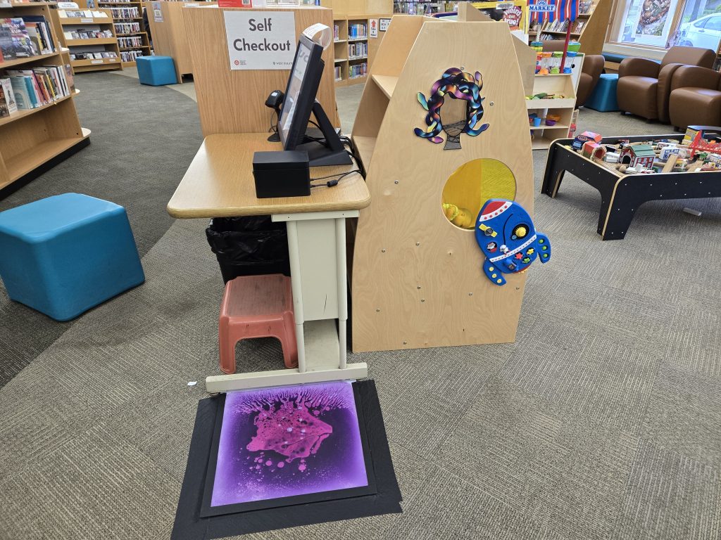 An image of a self checkout station next to a purple and pink liquid floor tile. 