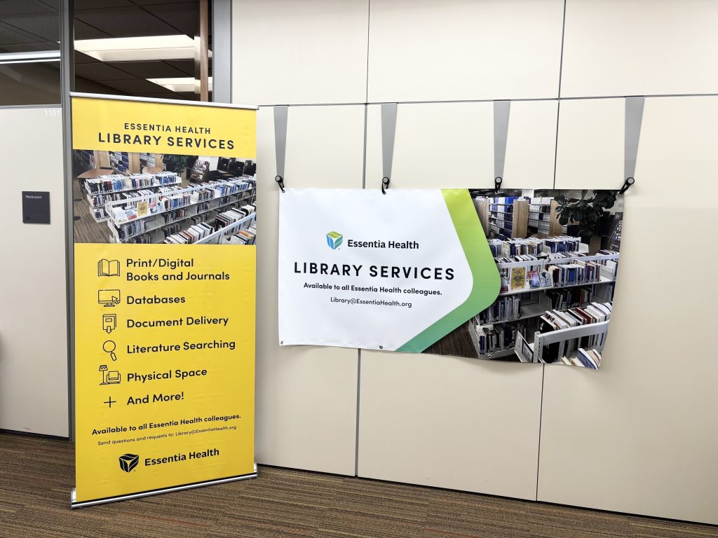Image shows one horizontal Essentia Health Library Services banner, and a vertical free standing yellow banner with listed services: Print/Digital Books and Journals, Databases, Document Delivery, Literature Searching, Physical Space and more! The services listed are "available to all Essentia Health colleagues".